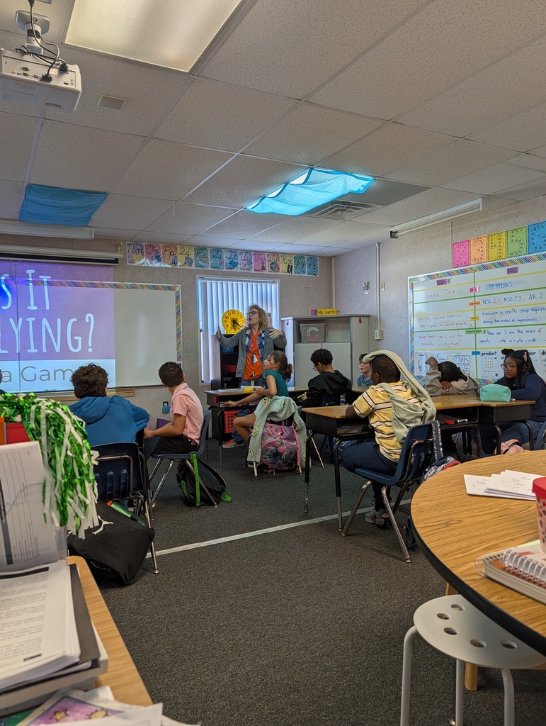 A classroom with students seated at tables. A teacher is standing at a projector screen displaying text.