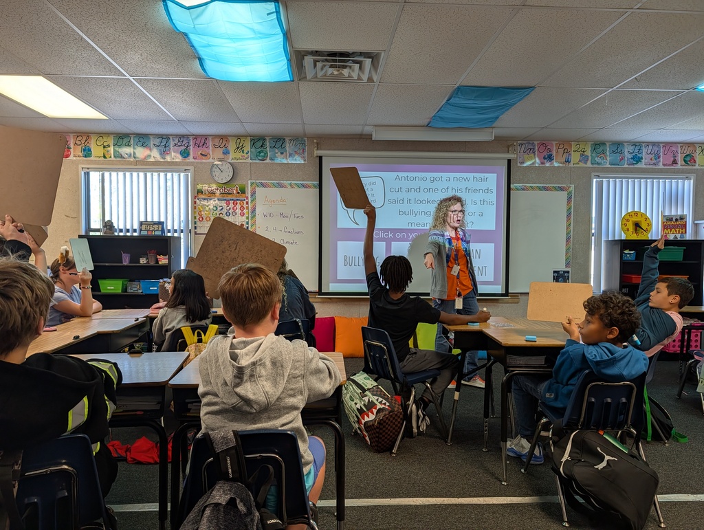 Students seated at desks in a classroom raise hands; a person stands at a podium, presenting.