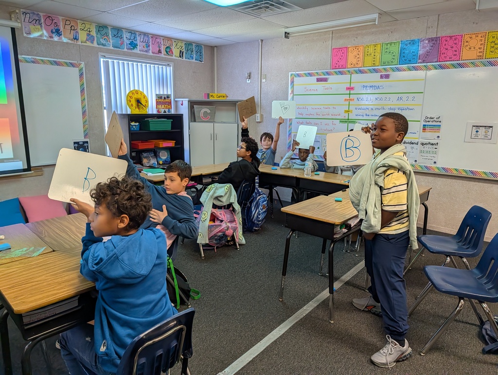 Classroom with desks and chairs. Students hold up paper with letters. Whiteboards and posters on wall.
