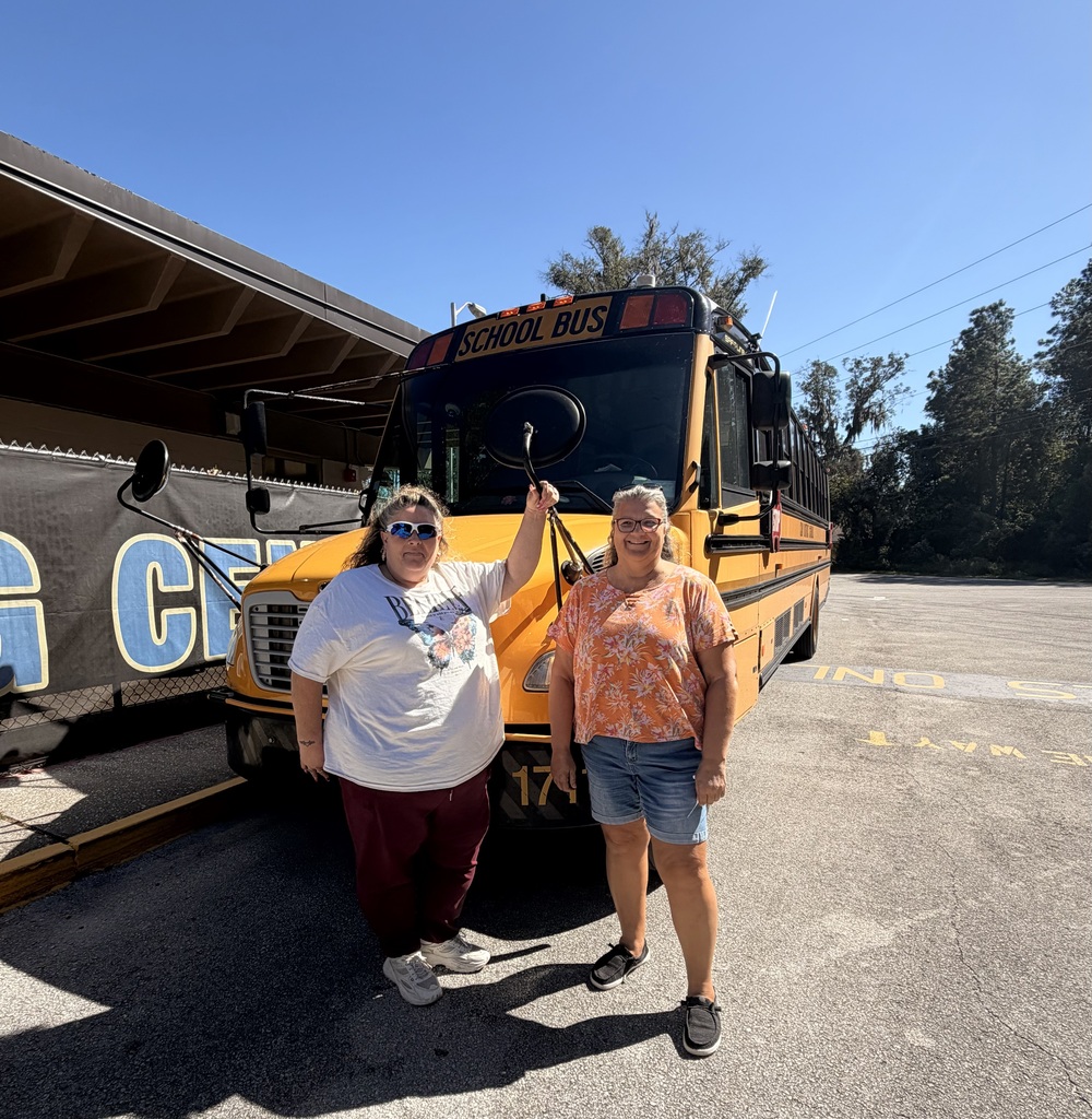 Two women stand in front of a parked yellow school bus. They hold up their hands.