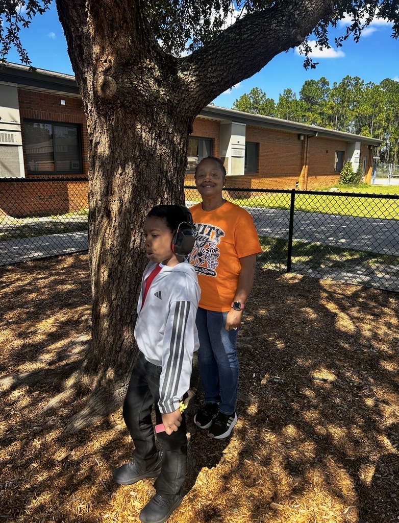 Two kids standing near tree in front of building with a chain-link fence.