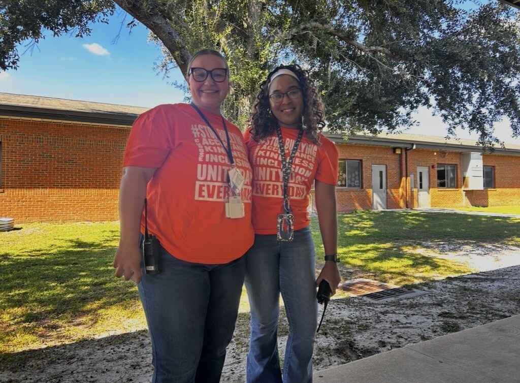 Two adults in orange t-shirts pose in front of a brick building with a tree.