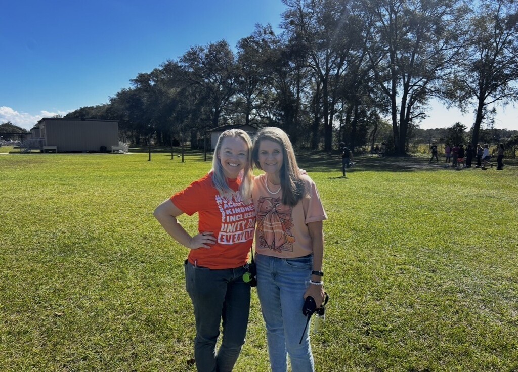 Two women standing in a grassy field with trees and a building in the background.