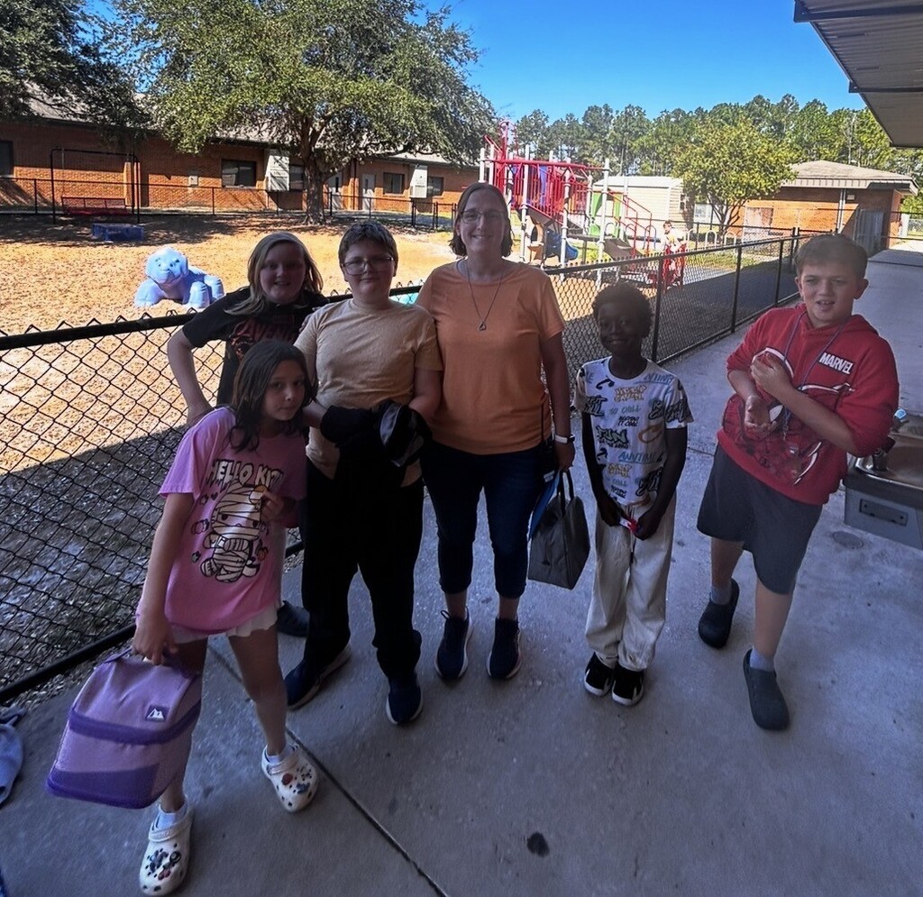 Adults and children stand in front of a fence with playground equipment behind it.