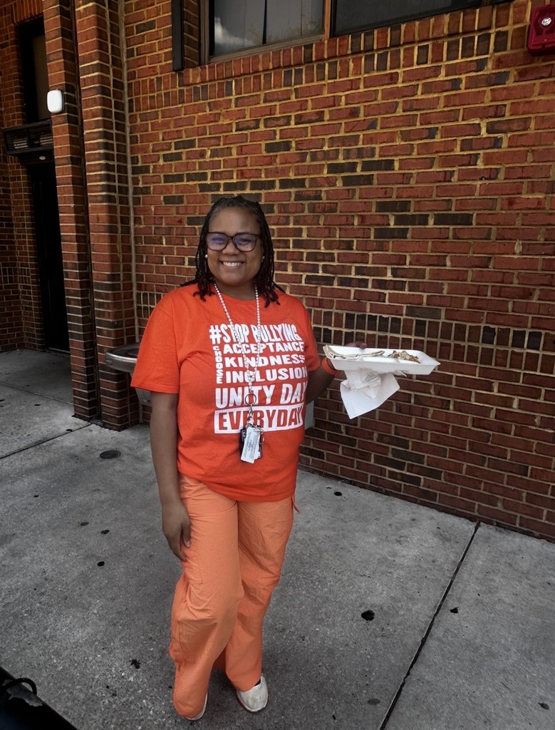 Person in orange shirt and pants holds food on tray in front of brick wall.