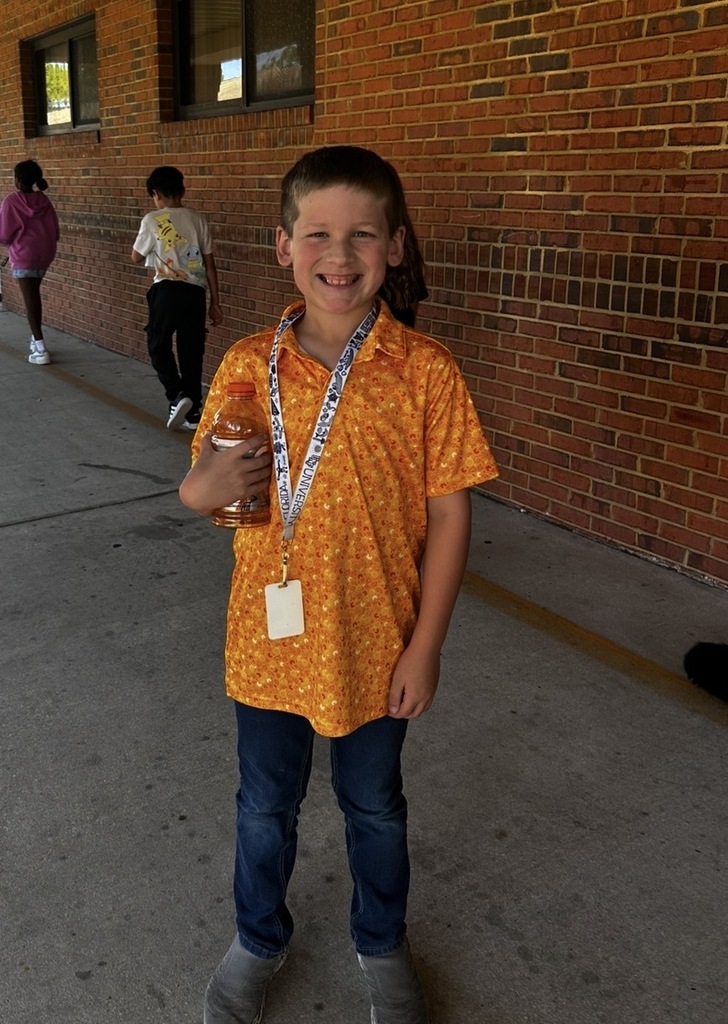 A young boy smiles in front of a brick wall; he wears a lanyard and holds a drink.