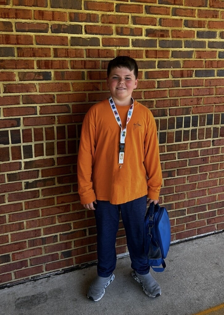 A boy in orange shirt and blue pants stands next to a brick wall, holding a blue backpack.