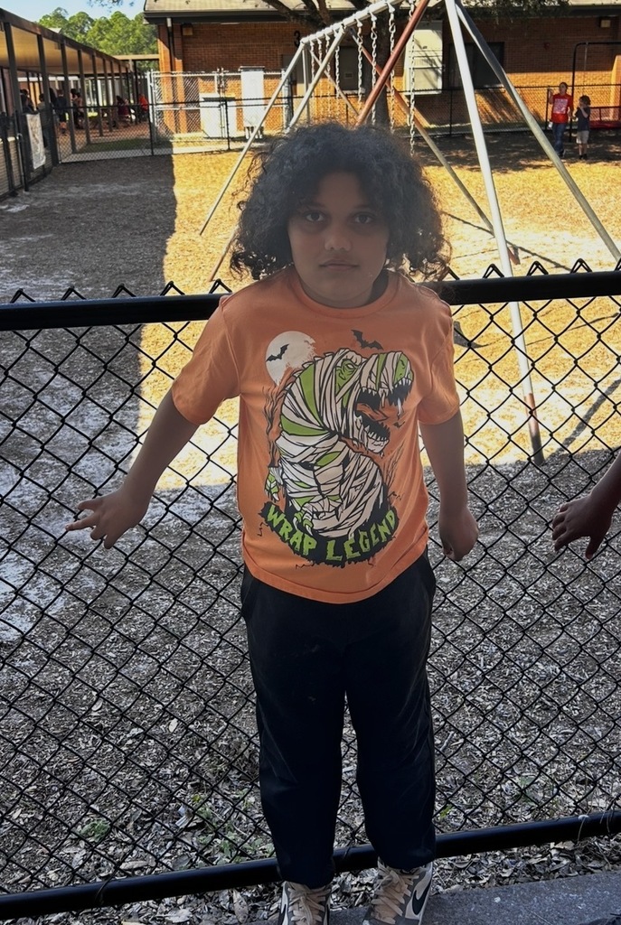 A child in a bright orange shirt with a Halloween graphic stands by a black fence.
