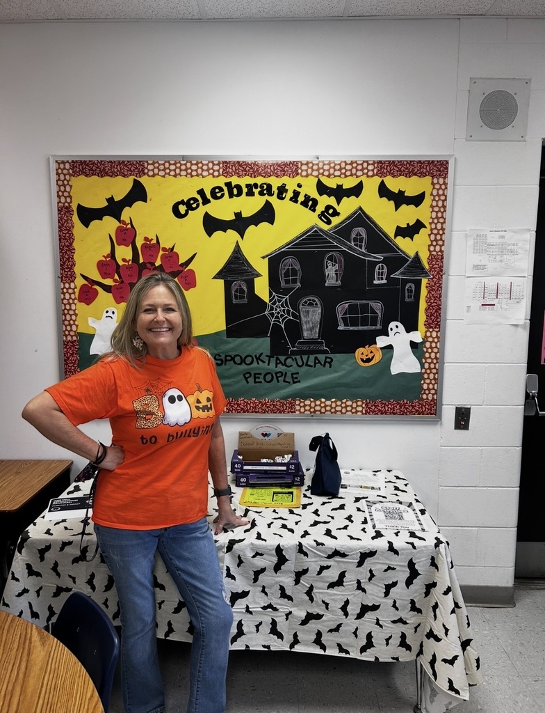 Woman in orange shirt stands in front of a table with Halloween decorations, near a Halloween poster.