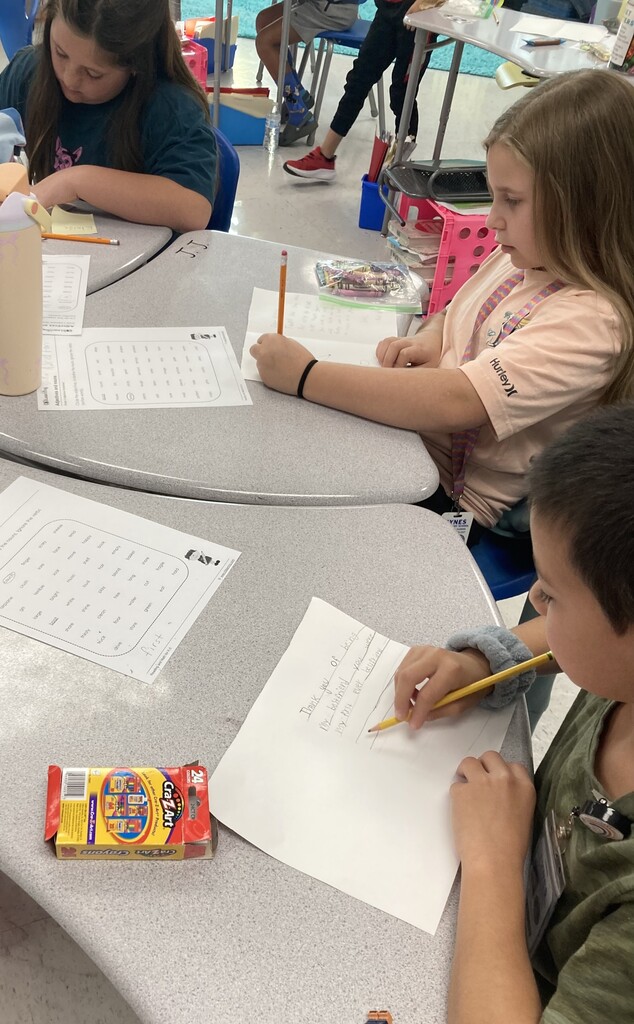 Four children at desks, each with writing utensils, papers, and a box of crayons. One child is writing.