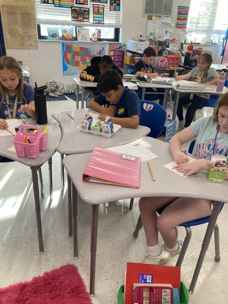 Students in a classroom sit at desks, working on papers. Colored pencils, pens, and folders are on the desks.