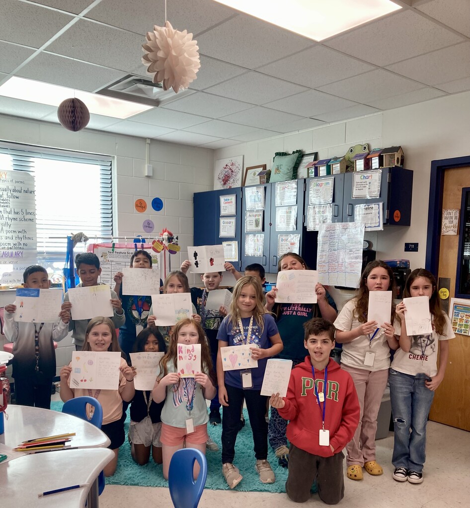 Children in a classroom pose with white papers; some are holding. A desk, chairs, and a door are in the background.