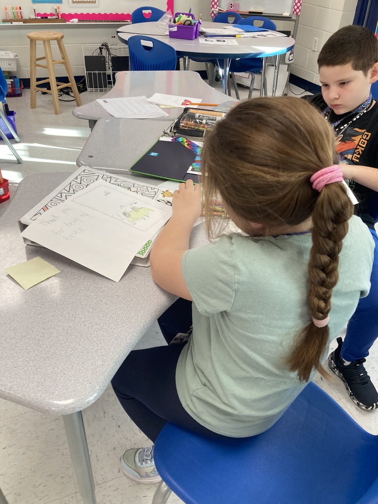 Children sitting at a table with papers and pencils. A boy sits behind a girl with a ponytail.