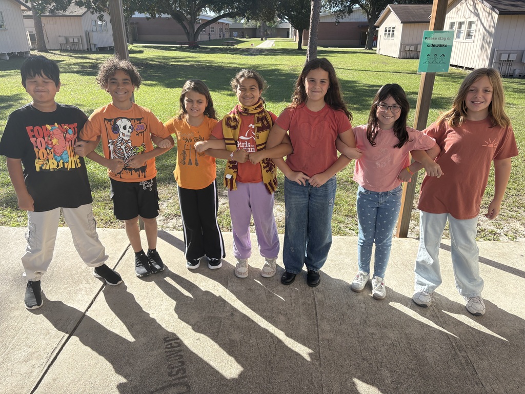 Eight children in matching tops stand on a sidewalk, arms linked, with a grassy area and houses in the background.