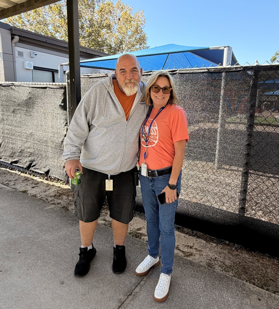 A man and a woman stand outside a fence with a building and trees behind.