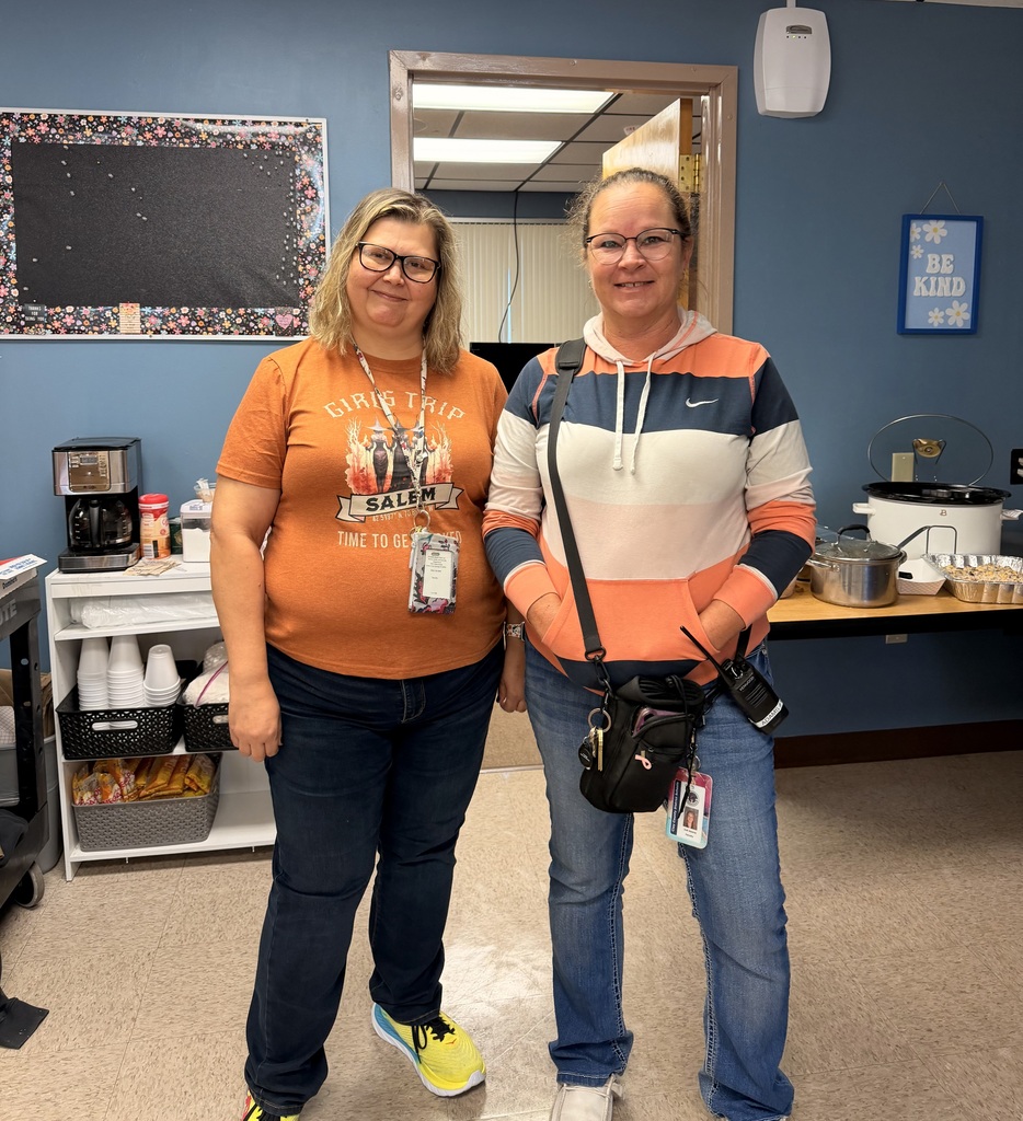 Two women stand in a room with blue walls and a white shelf with coffee cups. One wears an orange shirt, the other an orange and white hoodie.