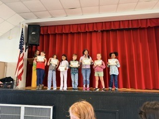 A group of young children stand on stage holding certificates while an American flag stands behind them.