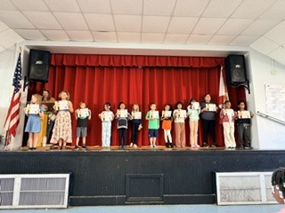 Children stand on stage with certificates, with a flag and red curtain in the background.