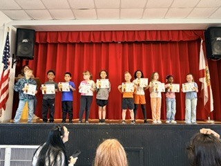 Students stand on a stage, holding certificates, with a red curtain and two flags behind them.