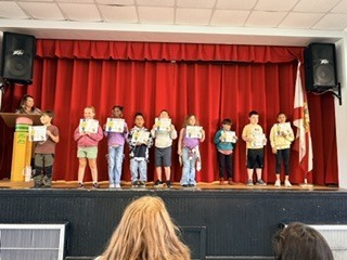 A group of children holding certificates on a stage with a red curtain. A woman speaks at a podium.