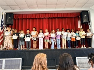 Several children holding certificates stand on stage under a red curtain; two flags are in the background.
