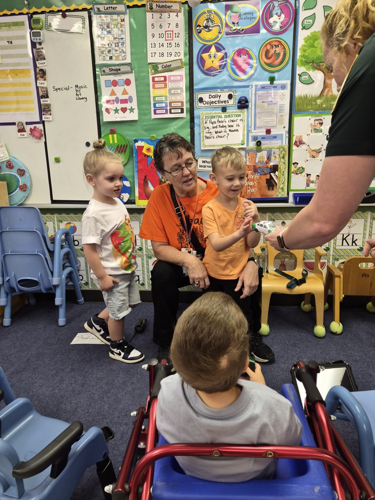 Woman sitting with three children, one in wheelchair, standing person holding item, and chairs in background.