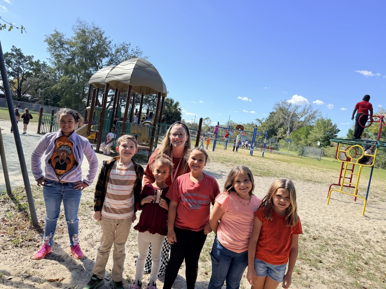 Several young girls and an adult woman in a schoolyard with a playground in the background.