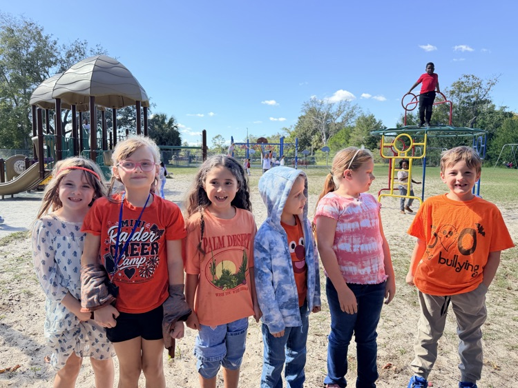 Several children in orange, pink, and blue shirts and pants stand in a playground with a slide.