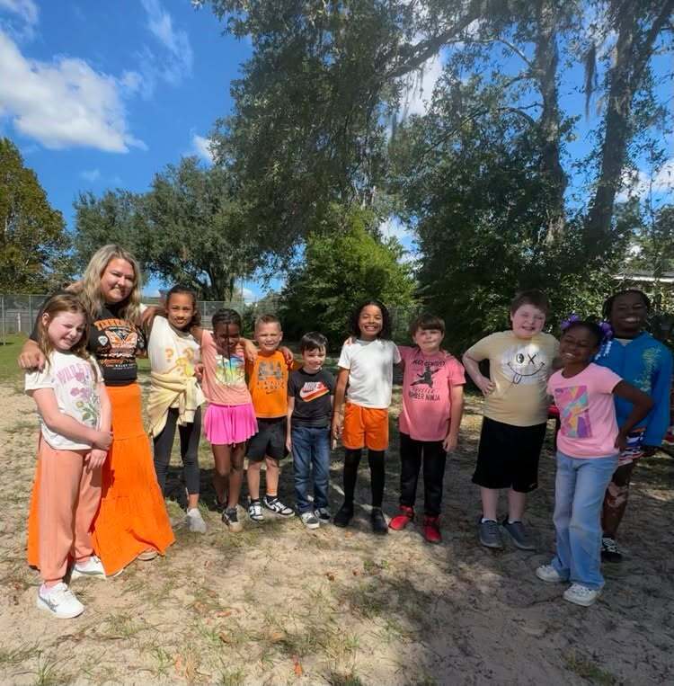 A group of children standing in dirt, a woman among them. Behind them are trees and a building.