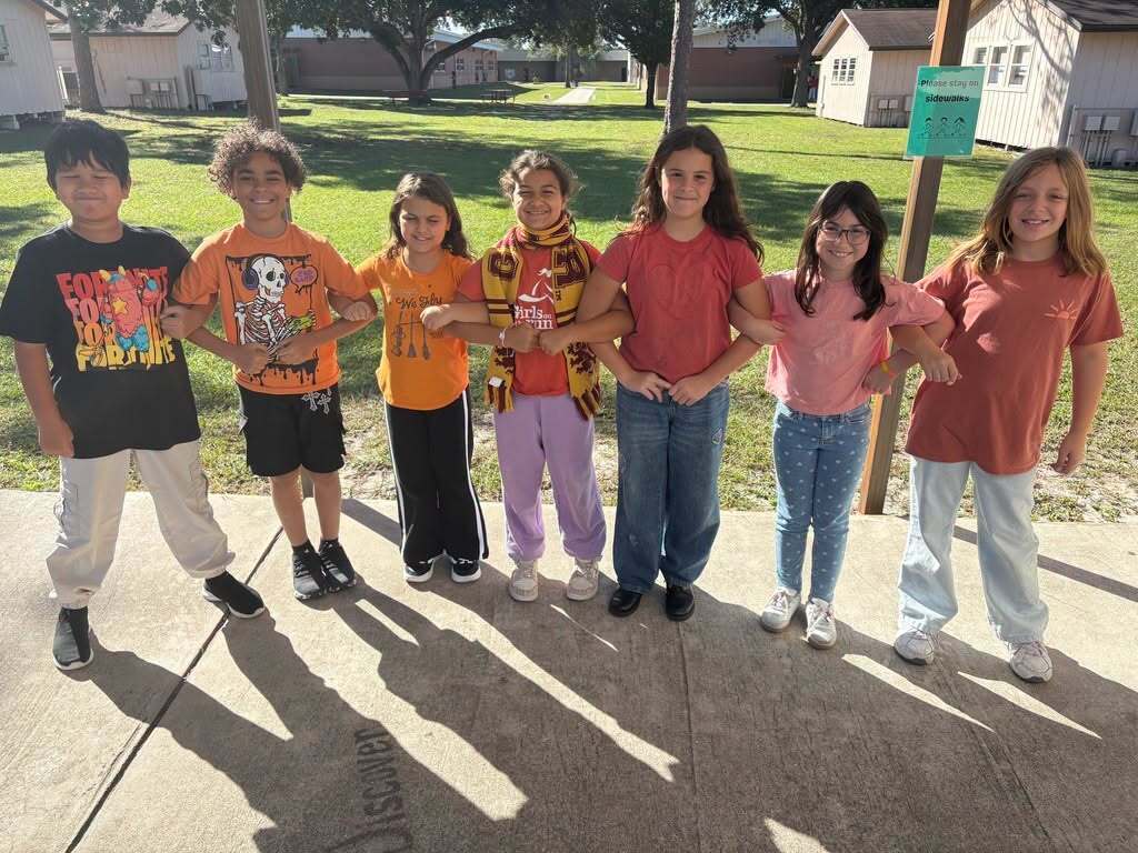 Nine children stand in a row, holding hands, in front of a grassy area. Behind them, a pole with a sign is visible.