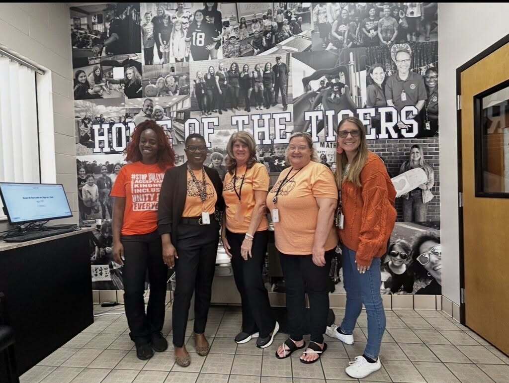 Five women stand in a room, smiling at camera. A large collage of photos on wall behind them.