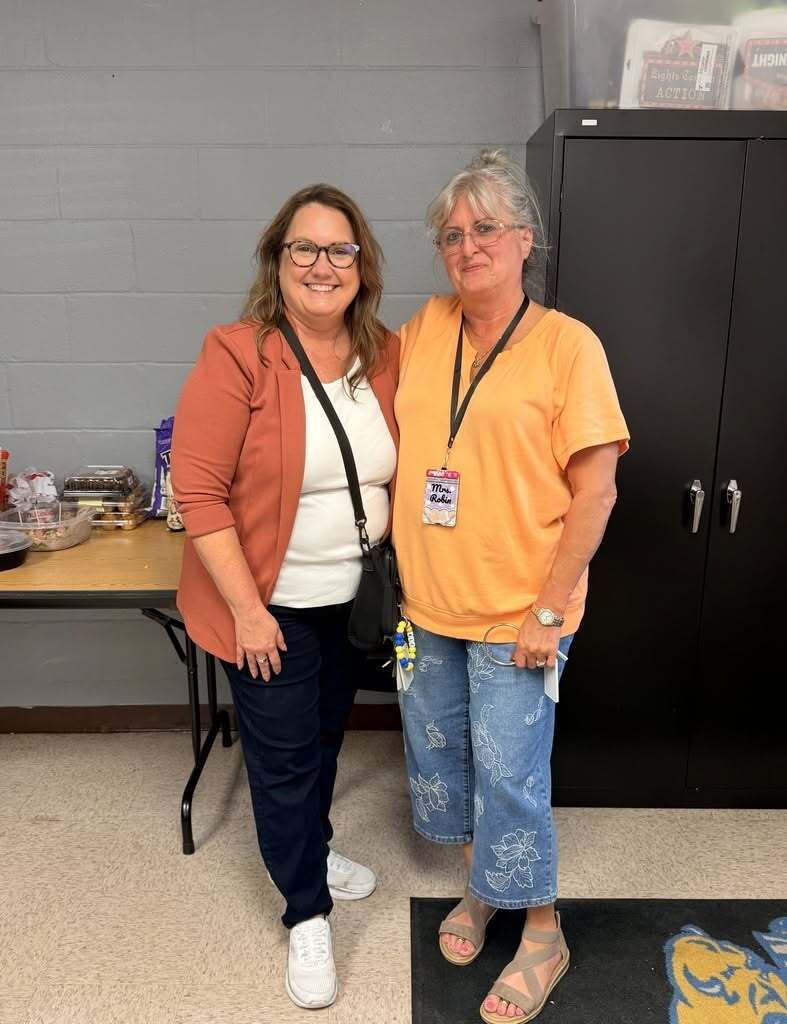 Two adults standing next to each other, one wearing a watch and the other wearing a backpack. Behind them is a black storage cabinet.