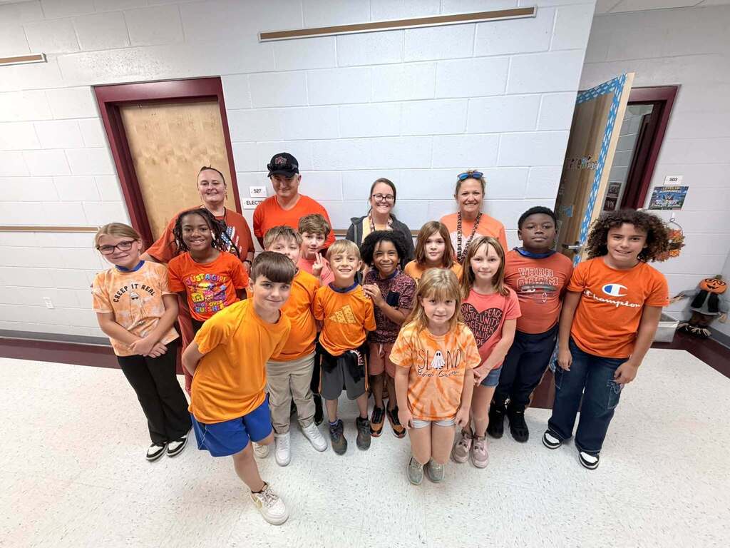 Group of young children in orange t-shirts and adults in a corridor, smiling for a photo.