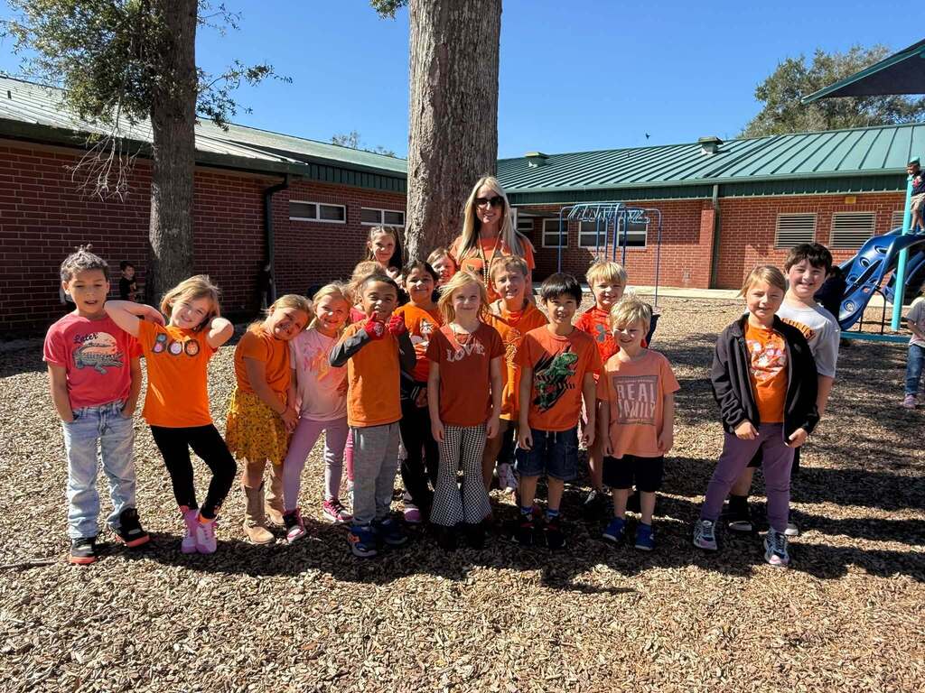 A group of children wearing orange shirts stands near a tree and a building with a green roof.