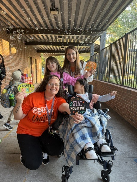 A woman kneeling and two children in wheelchairs holding signs; one sign says "STAY POSITIVE".