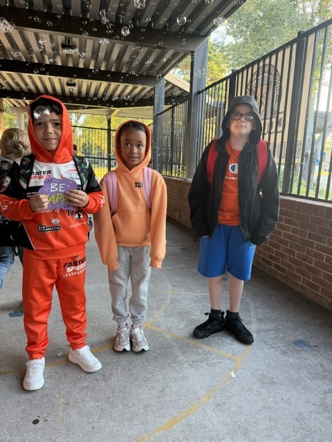 Three students stand in a covered outdoor area with brick walls. One holds a book, one wears glasses, and the third wears a backpack.