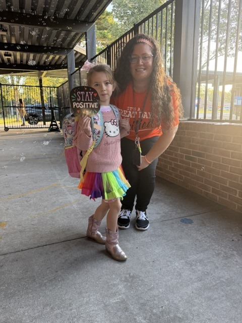 Woman in orange shirt and girl in pink shirt, rainbow skirt, and boots holding a backpack, posing with a sign.