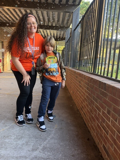 A woman and a child stand outside, wearing orange shirts and sneakers, near a brick wall and black railings.