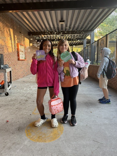 Two girls smiling at camera, holding items, one in pink jacket, one in orange shirt, standing in hallway, another person with backpack nearby.