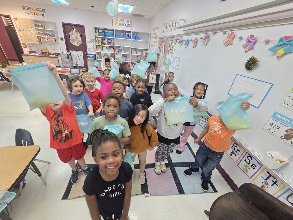 A classroom with children holding up clear bags. The walls are decorated with artwork and shelves with books.