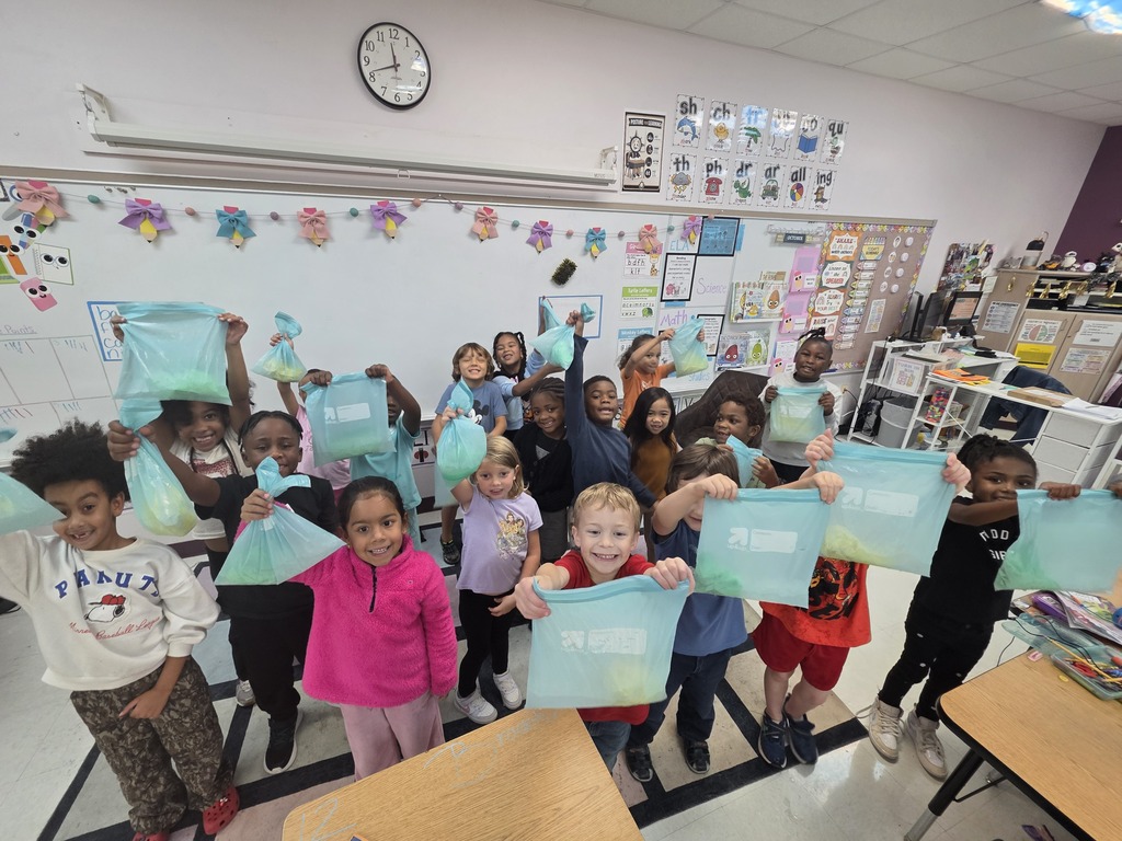 Children holding up bags in a classroom with whiteboard, bulletin board, clock, and tables.