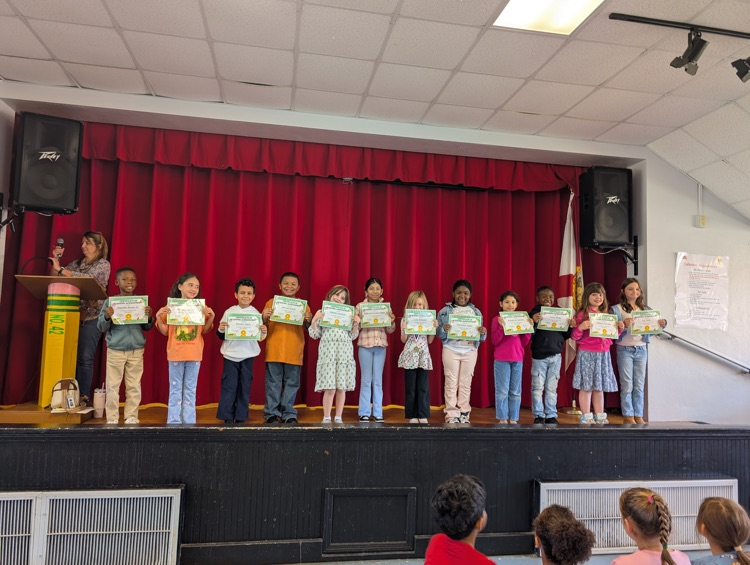 Children standing in two rows on stage holding certificates, one adult at a podium.