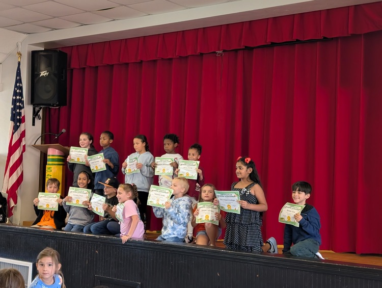 A line of young children on stage holding certificates in front of a red curtain.