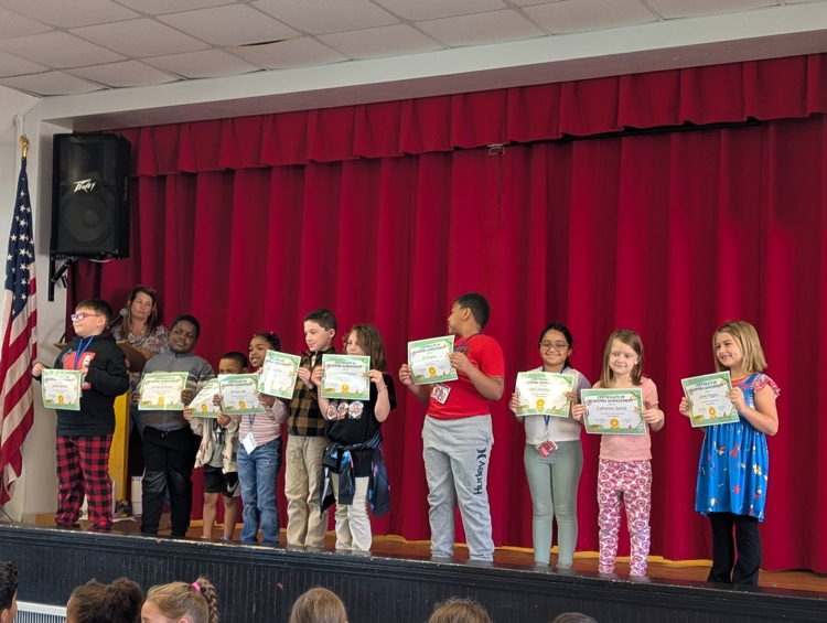 Several students holding certificates stand on stage in front of a red curtain, with an American flag on the left.