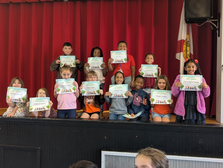 Group of young students holding certificates, standing on stage, under red curtains. One student holds a flag.