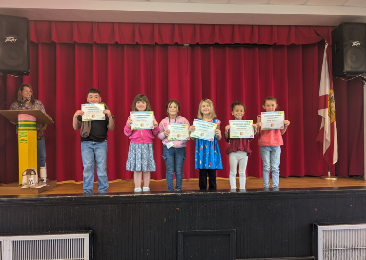 Students stand on stage holding certificates in front of a red curtain, with a flag and two speakers.