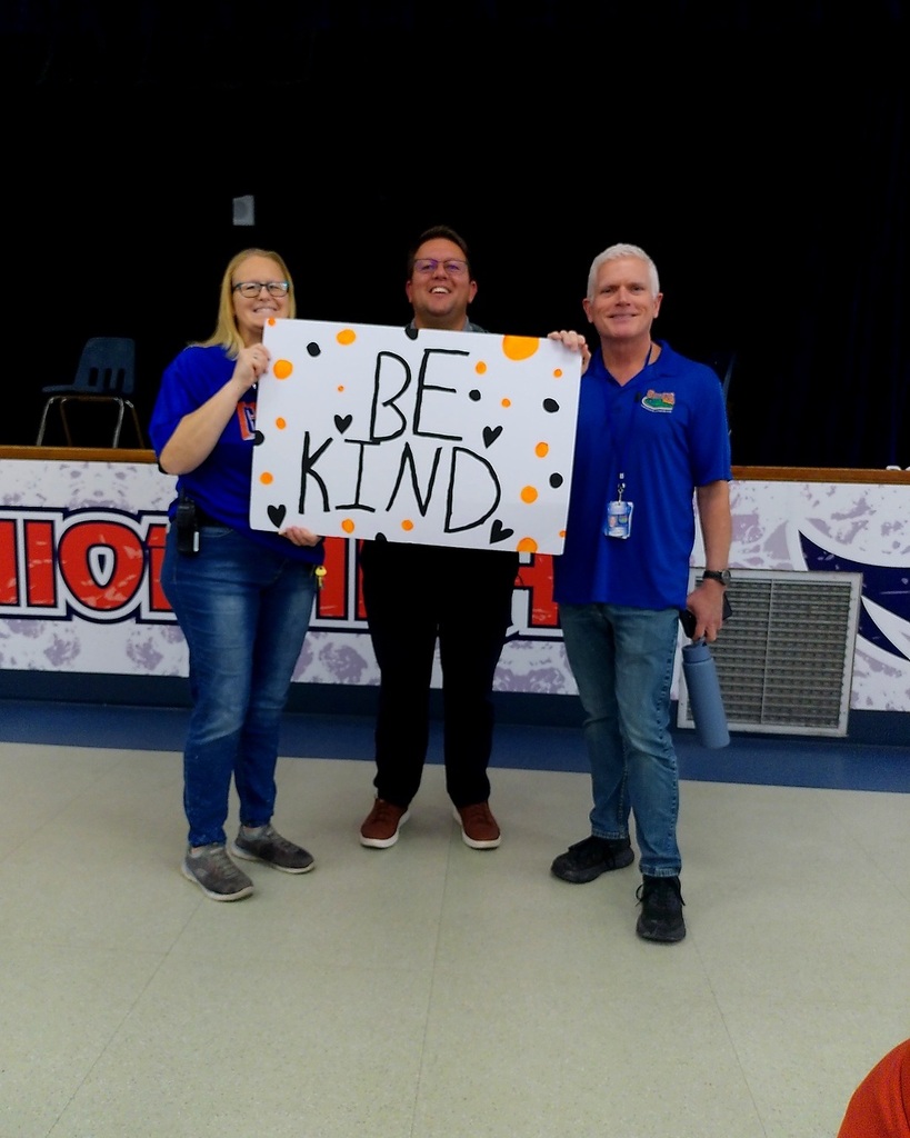 Three adults stand holding a white sign with orange polka dots. The sign says "BE KIND." Behind them is a white wall with orange letters.