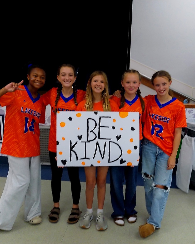 Five girls pose with a sign that reads, "Be Kind" while standing next to each other.