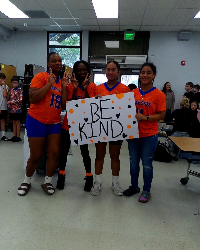 Four young women in orange shirts hold a white sign that reads "BE KIND". One holds a pumpkin.