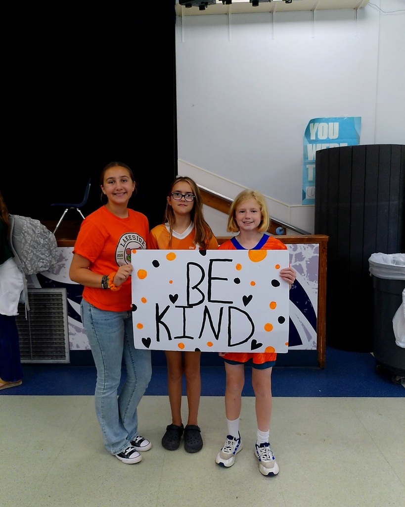 Three young girls hold a white poster with orange and black dots and black text reading 'Be Kind'.